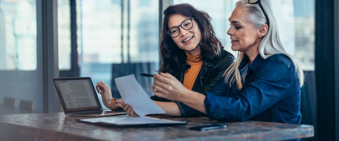 Women in discussion: one with a laptop, the other with papers